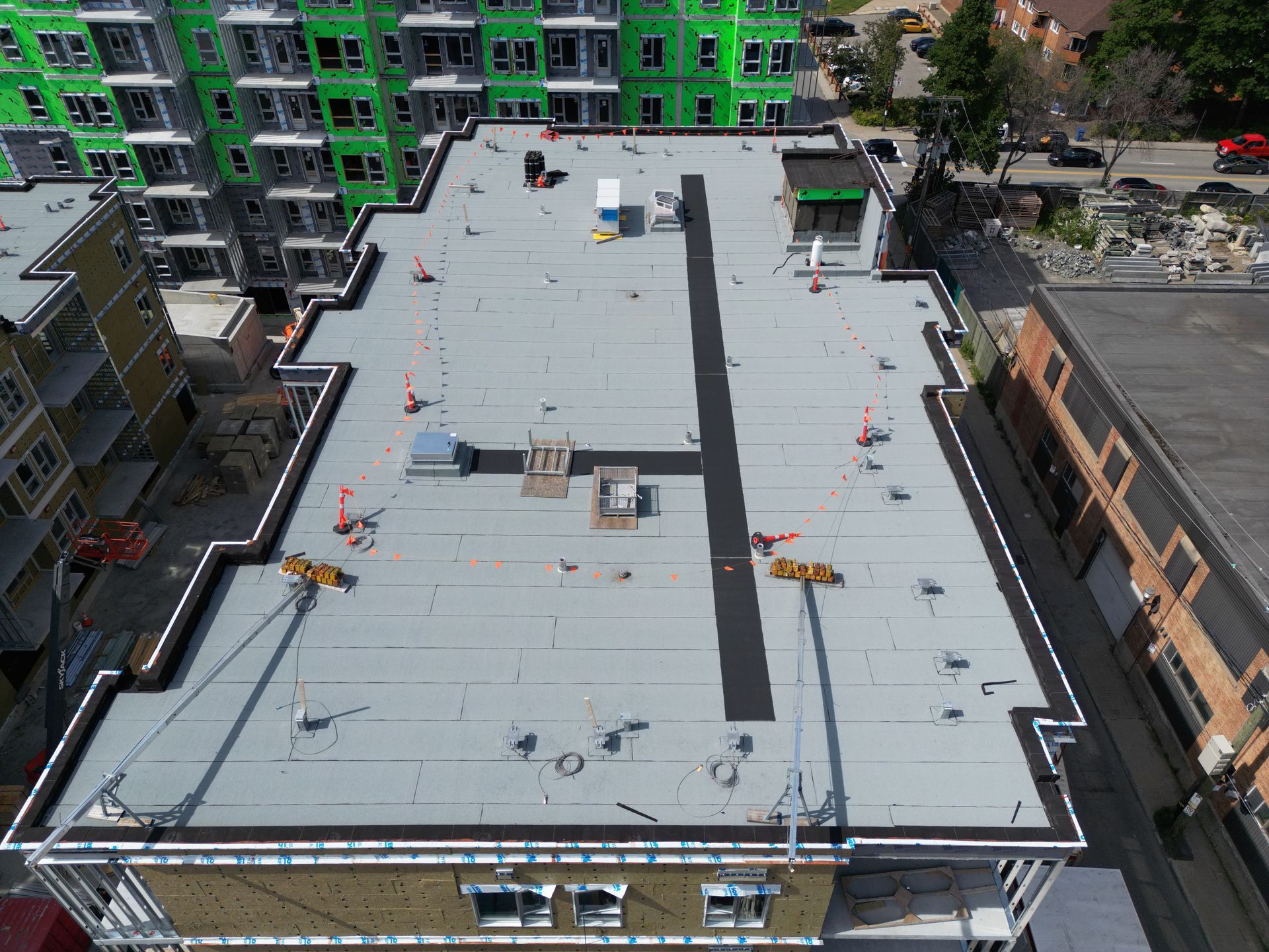 Rooftop construction site on a large flat building, with workers, equipment, and surrounding city buildings.