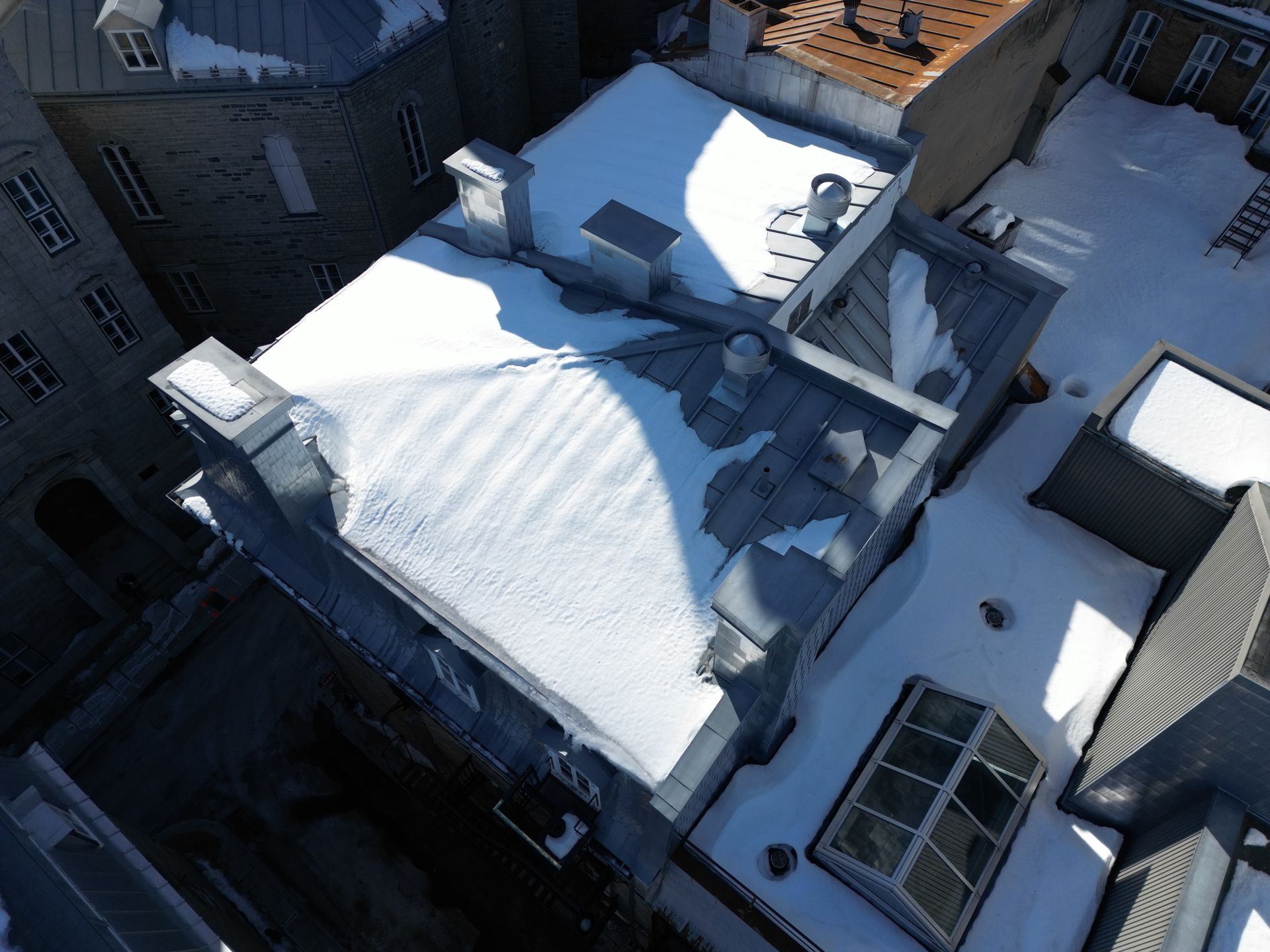 Snow-covered urban rooftops viewed from above, with skylights, vents, and adjacent brick buildings.