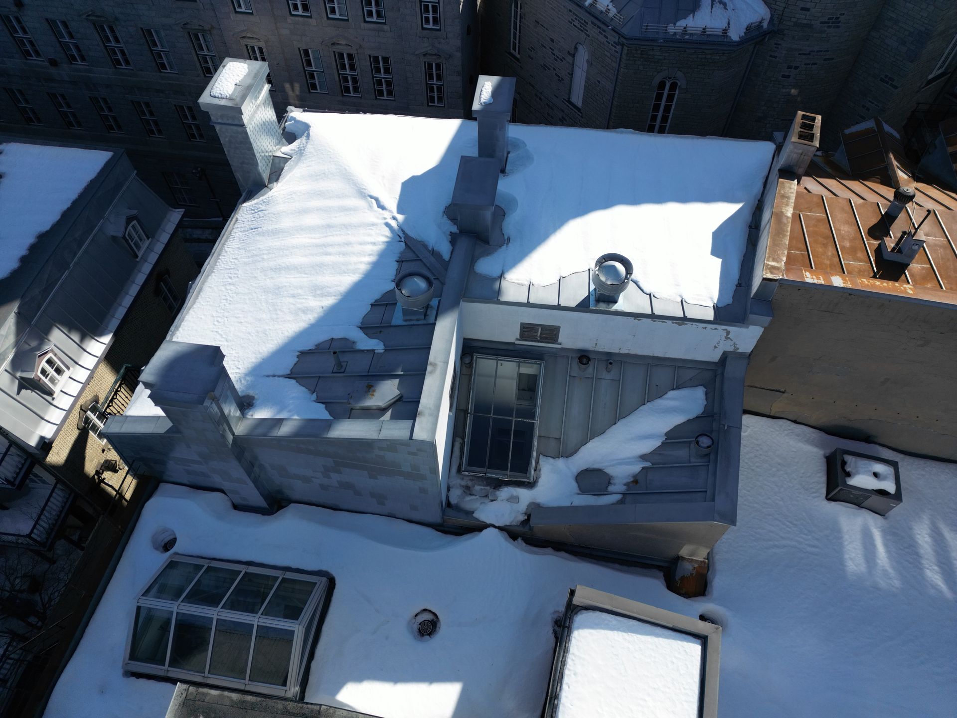 Snow-covered rooftops beside a brick building, viewed from above in bright daylight.
