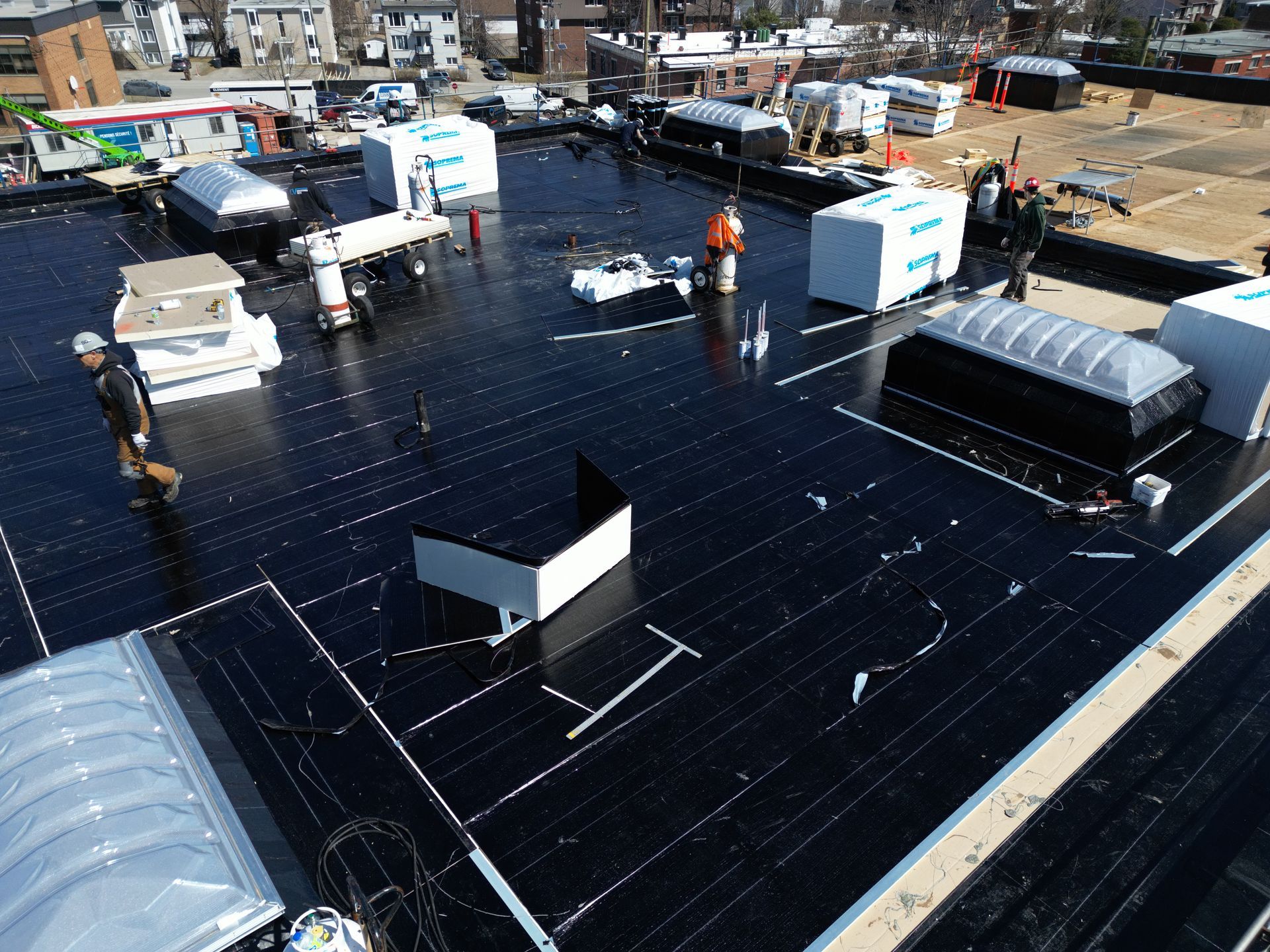 Rooftop under construction with workers, black roofing material, and HVAC units in an urban area