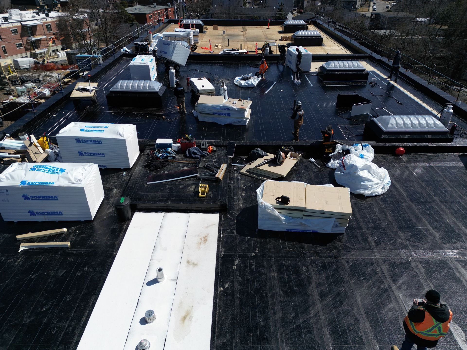 Rooftop under construction with HVAC units, workers, and dark waterproofing material spread across the roof.