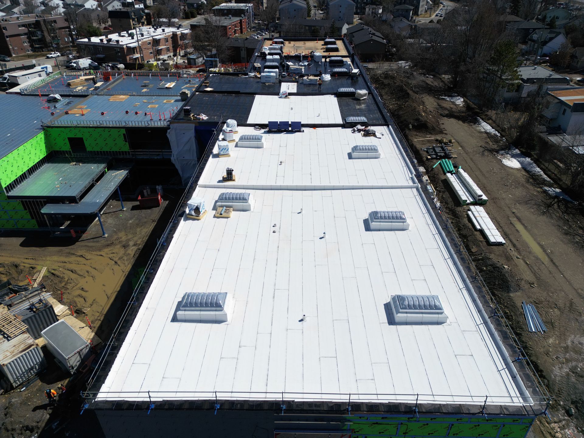 Aerial view of a large white-roofed building with rooftop vents and surrounding streets and trees.
