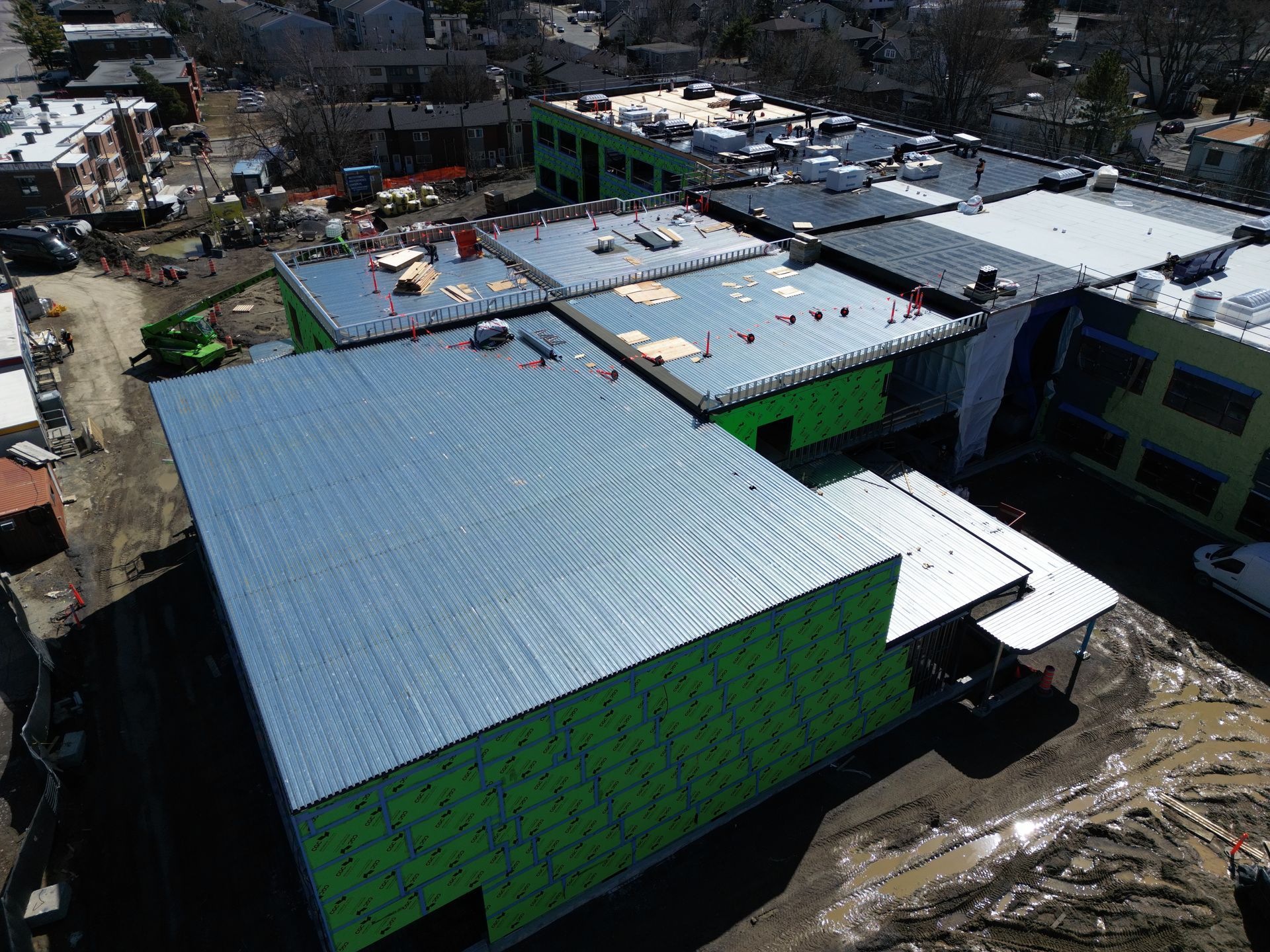 Aerial view of a large green building with a white roof beside other buildings and a muddy lot