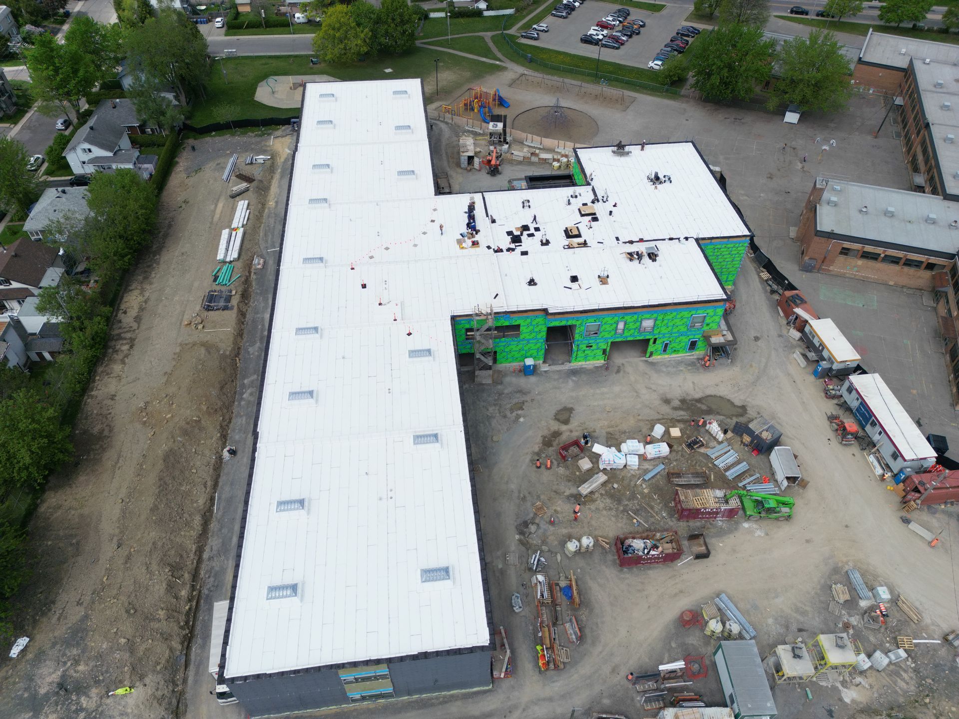 Aerial view of a large white-roofed building under construction with green exterior panels and nearby equipment.