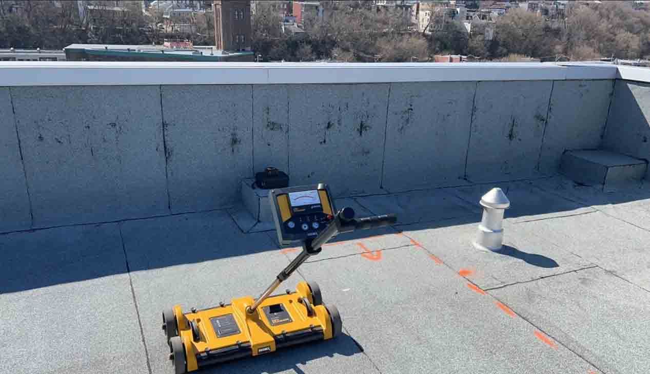 Un robot de chantier jaune se trouve sur un toit plat, à côté d'un muret et de bouches d'aération, avec un paysage urbain en arrière-plan.