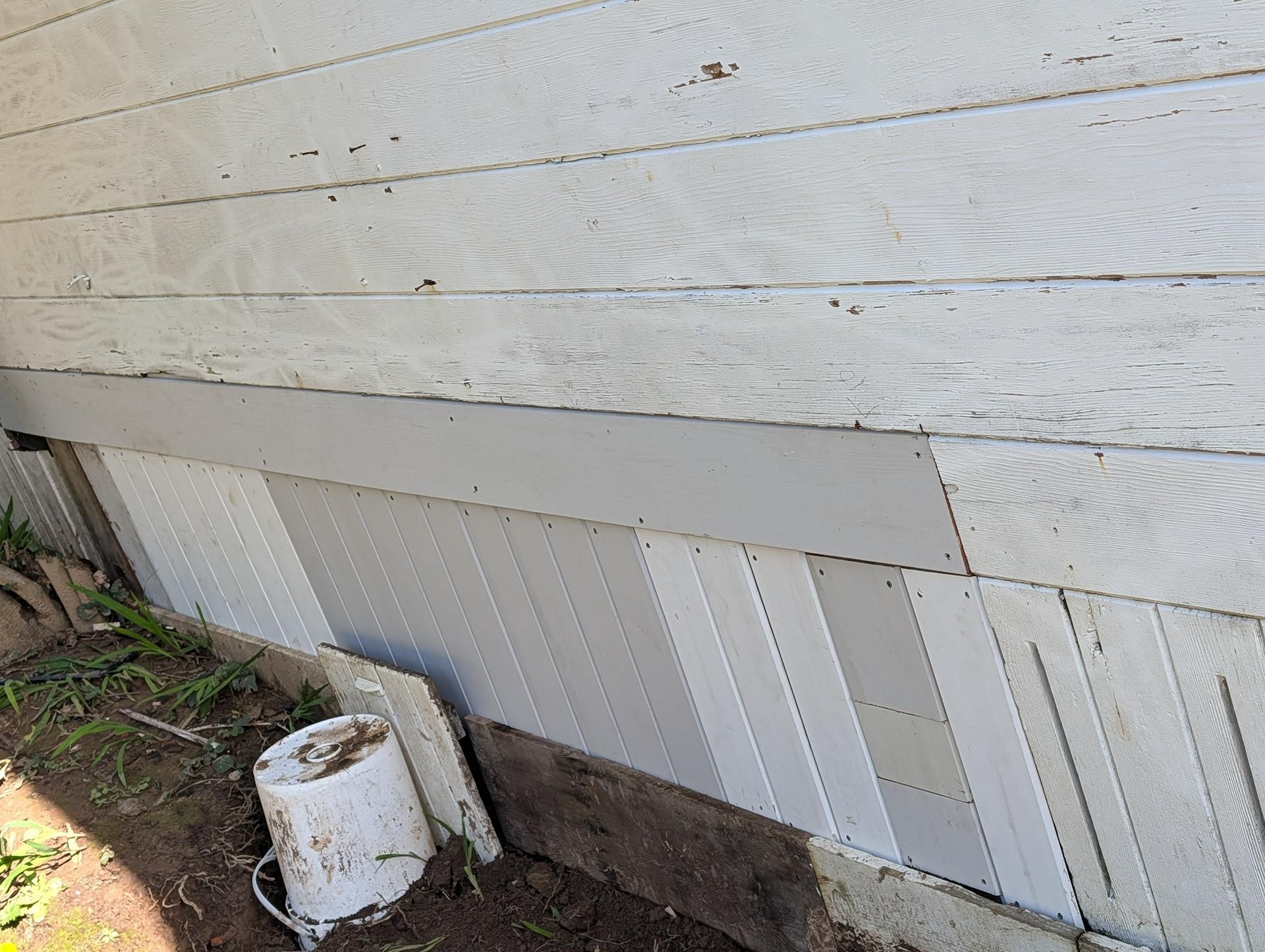 Damaged white exterior wall with peeling panels and a bucket on the ground near the foundation.