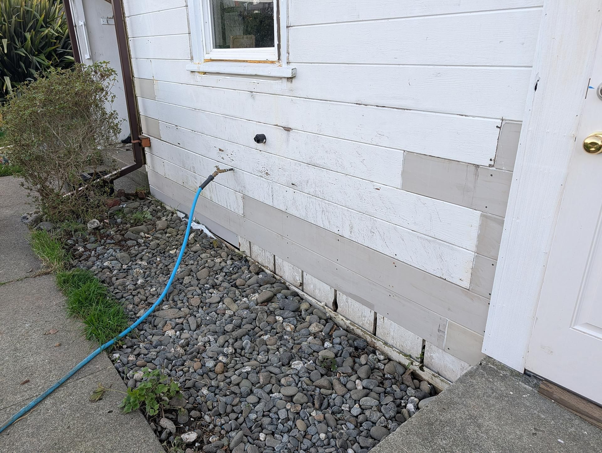 Exterior wall with patchy siding, a blue hose, and gravel along the base near a window and door.