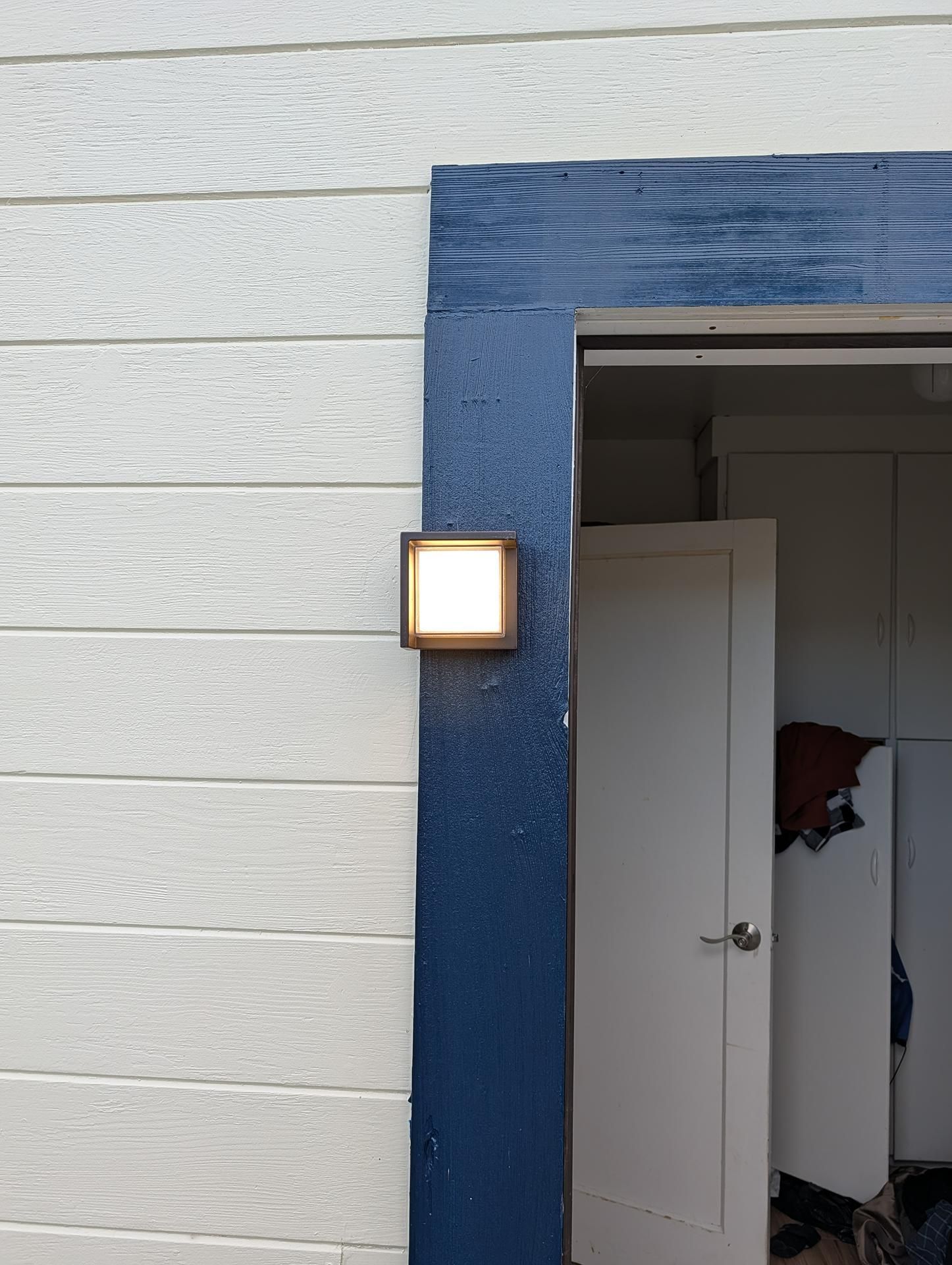 Blue doorway with a lit wall lamp beside an open white door revealing an interior room.