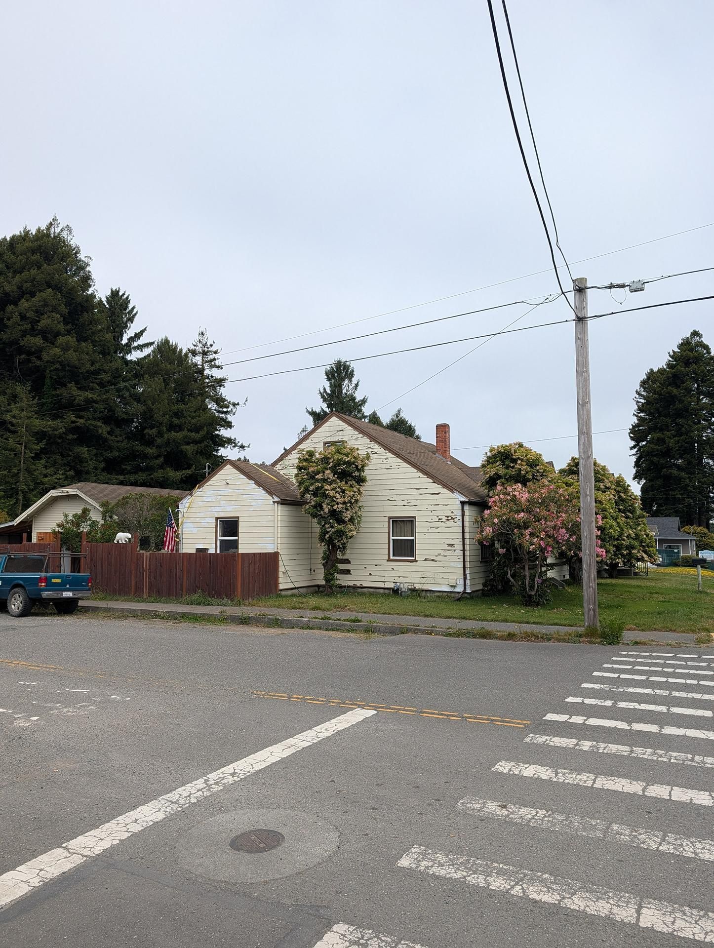 Small white house by a street corner with power lines, trees, and a parked blue truck.