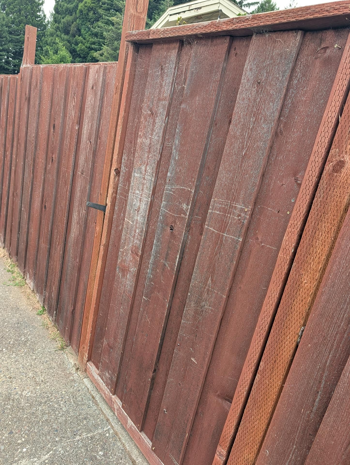 Rusty wooden fence lining a sidewalk, with greenery visible above it.