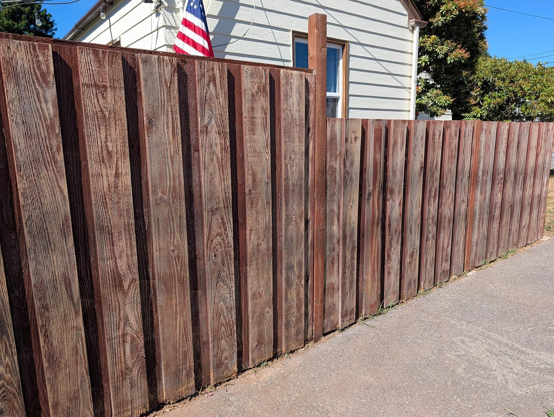 Tall weathered wooden fence along a driveway beside a house with a small American flag above it
