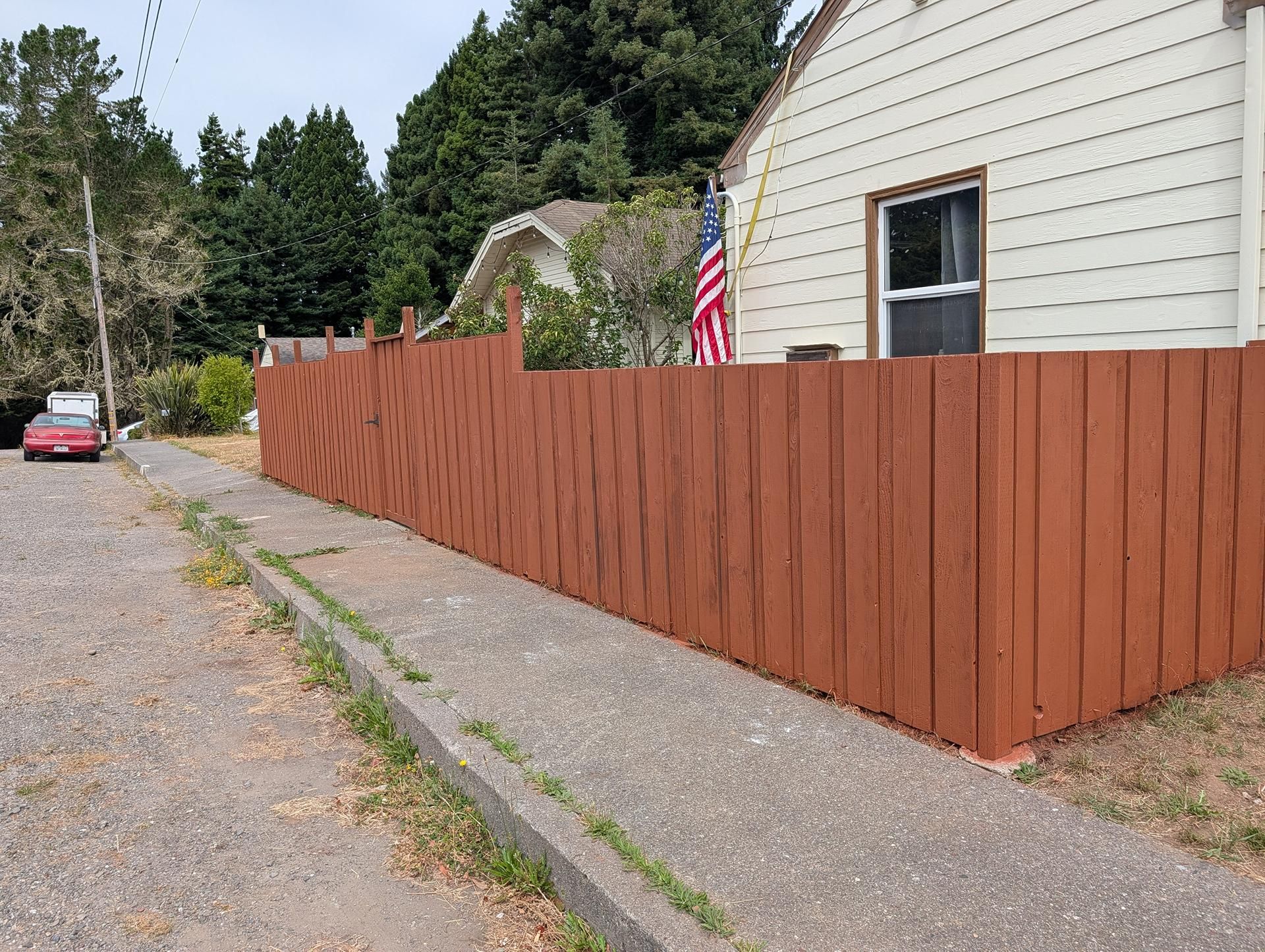Wooden fence along a driveway beside a white house, with an American flag and parked car in the background.