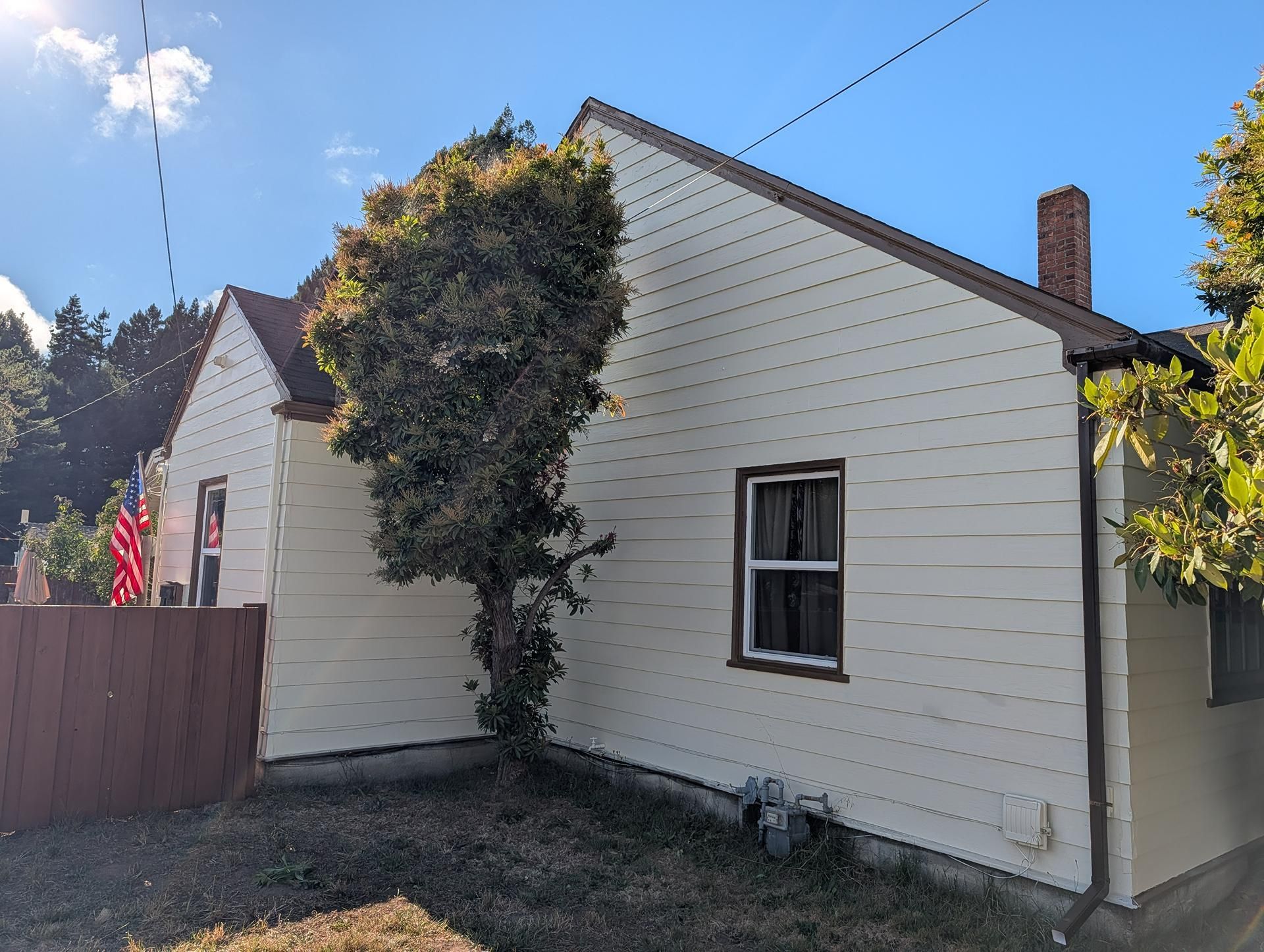 White house exterior with a small window, tree in front, and blue sky above the fenced yard