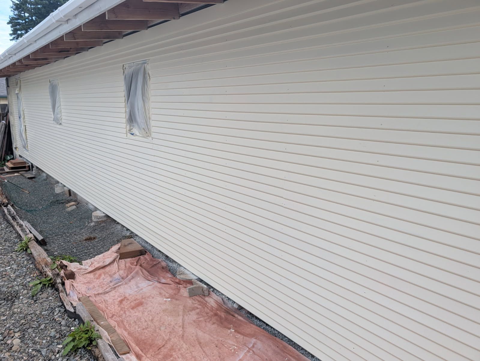 Side of a house under construction with white siding, covered windows, and pink insulation on the ground
