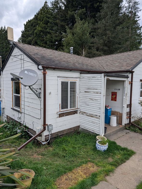 Small white weathered garden shed with open door, window, satellite dish, and surrounding grass.