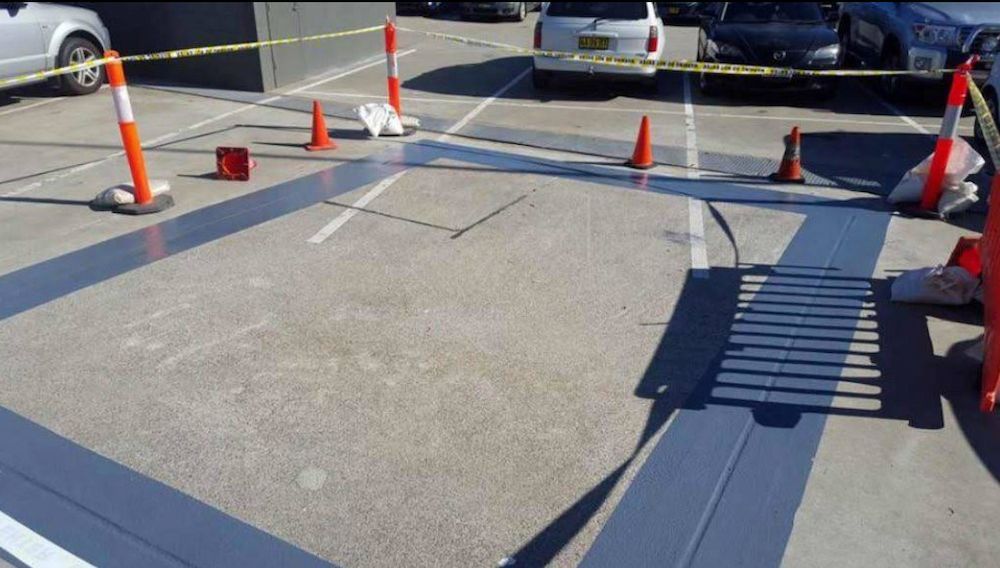 Parking space being renovated with fresh grey paint. Orange cones and caution tape surround the area with cars parked nearby — Consider It Sealed Waterproofing in Port Stephens, NSW