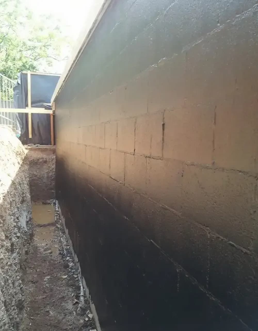 A foundation wall coated in black sealant, next to a narrow trench. The wall is brown brick — Consider it Sealed Waterproofing in Singleton, NSW