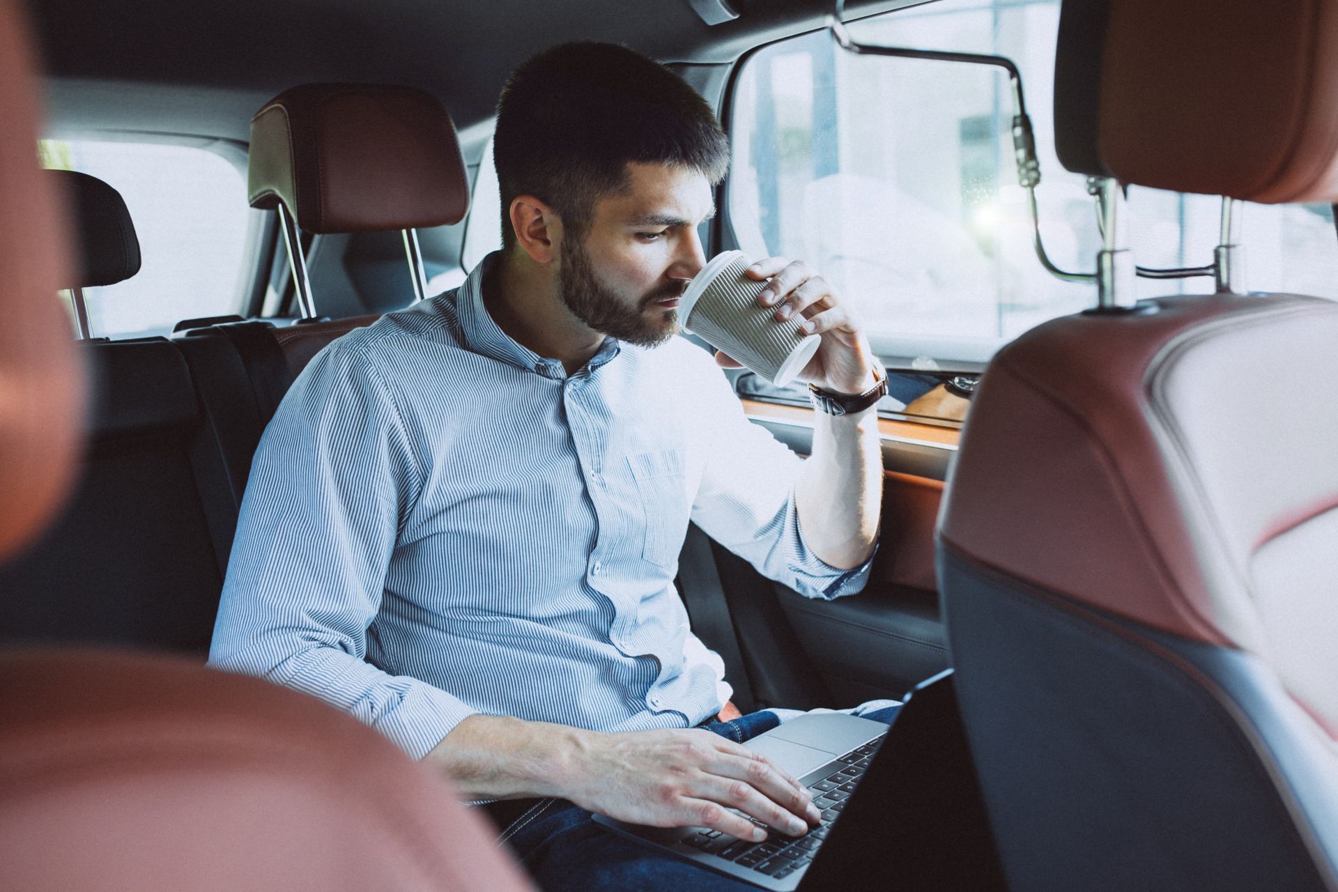 Hombre en un coche, tomando café y utilizando una computadora portátil en el asiento trasero.