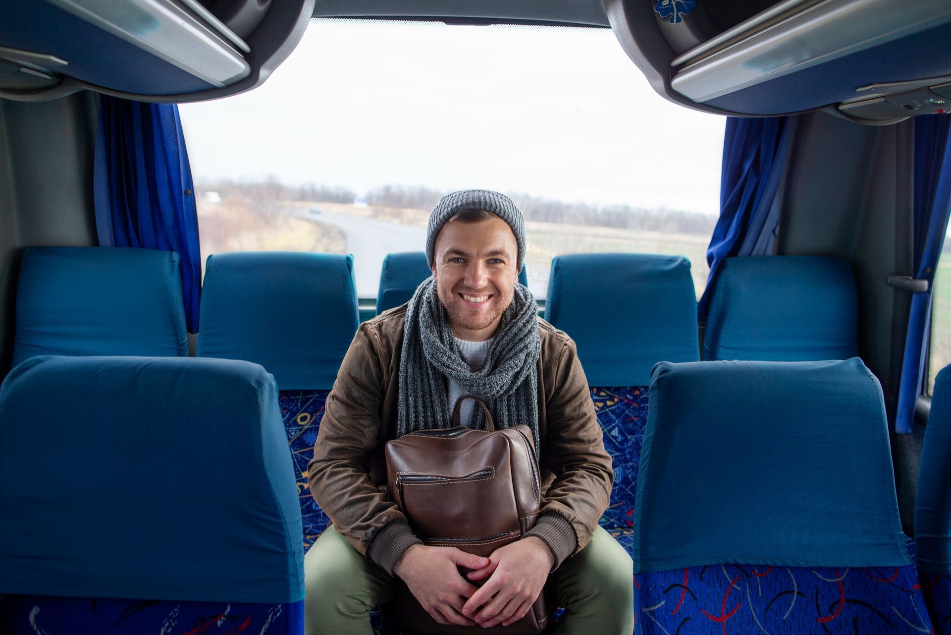Hombre sonriendo, sentado en un autobús con mochila. Lleva sombrero, bufanda y chaqueta. Vista interior.