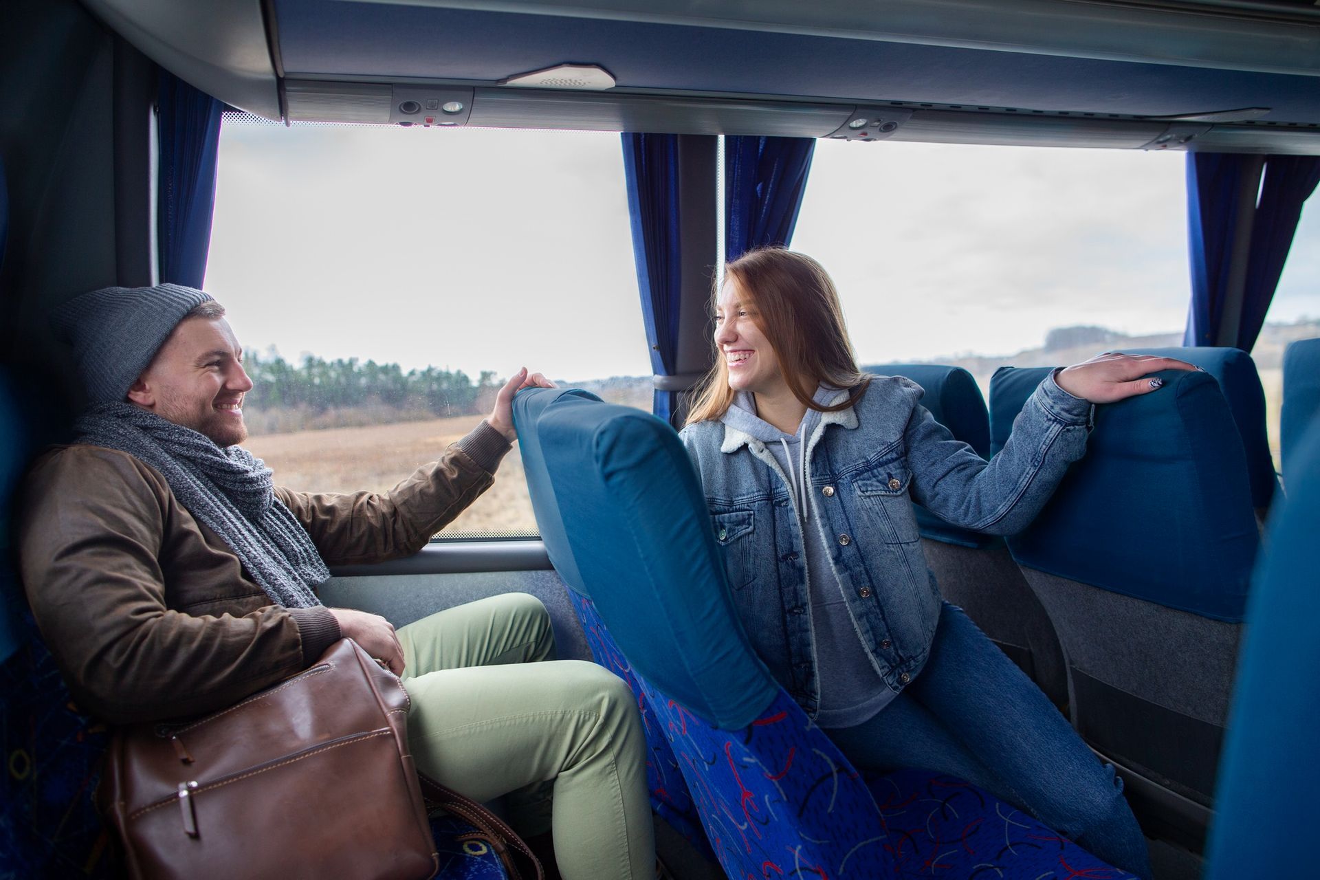 Pareja en un autobús, sonriendo, mirándose, con el brazo extendido. Fotografía interior con asientos azules y ventana.