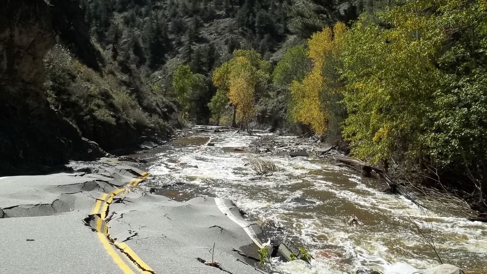 Damaged road washed out by floodwater in a wooded canyon, with debris and a broken yellow centerline