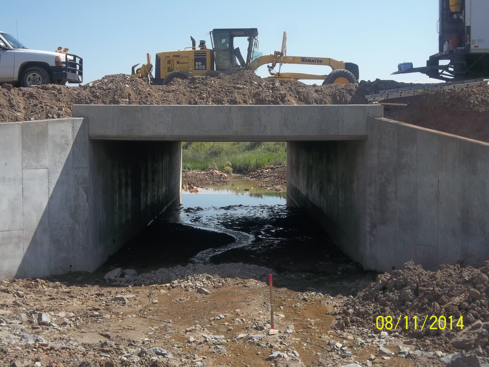 Concrete culvert under construction with excavator and dump truck on top, water flowing beneath.