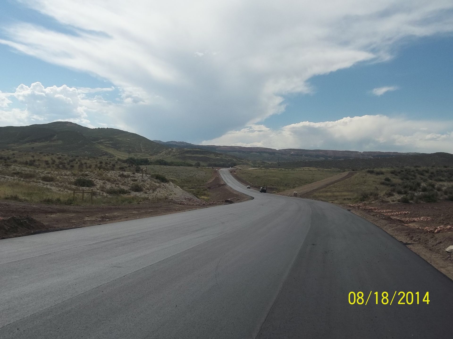 Rural paved road curving through hills under a cloudy sky, with date stamp in the corner