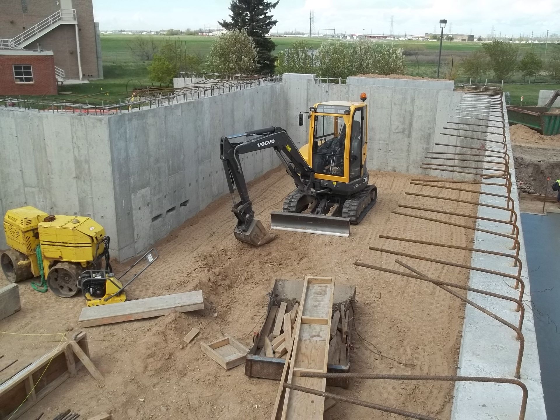 Excavator digging in a concrete foundation pit beside rebar and formwork at a construction site