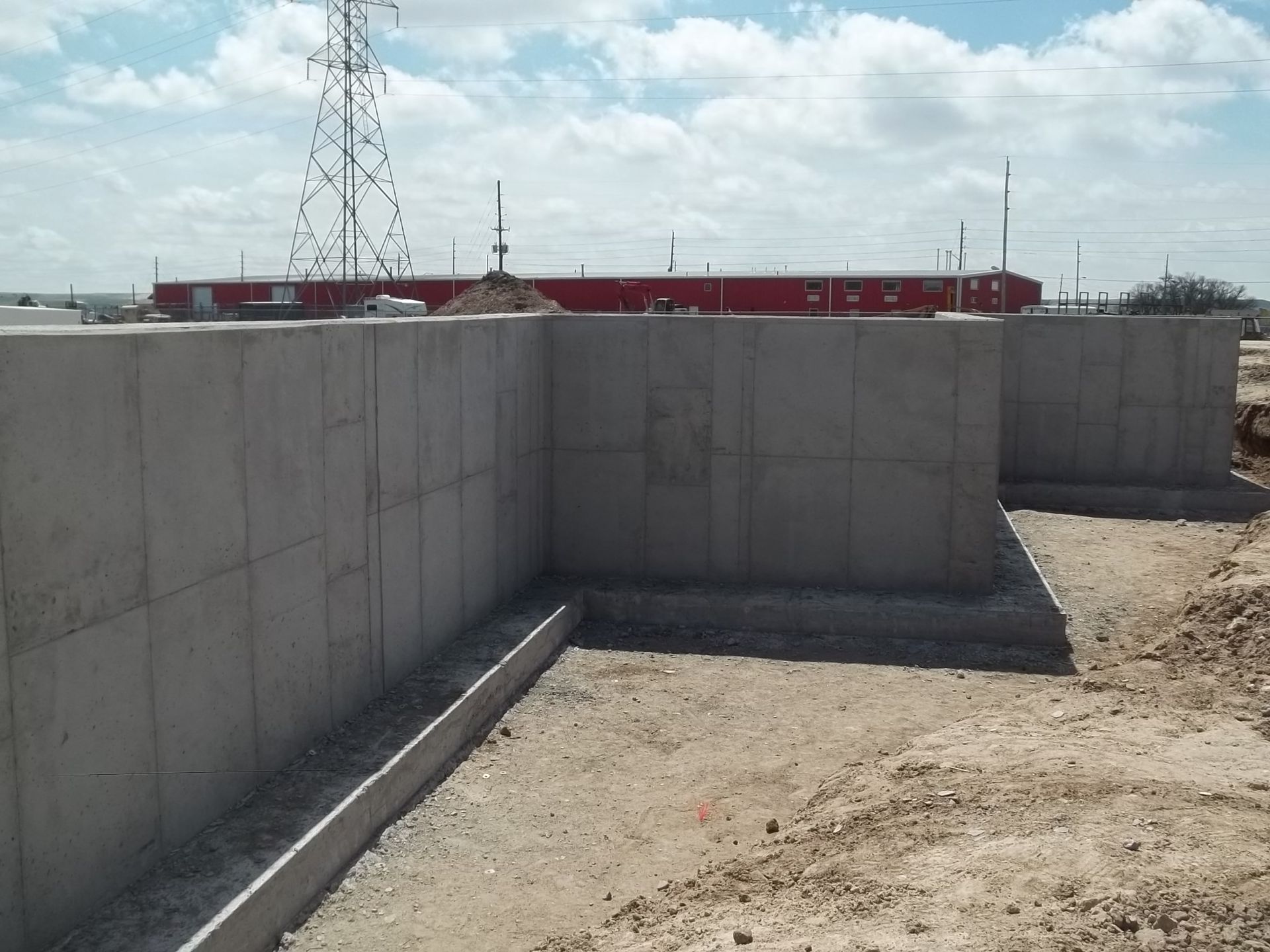 Concrete walls and dirt yard on a construction site with red buildings and utility towers in the background