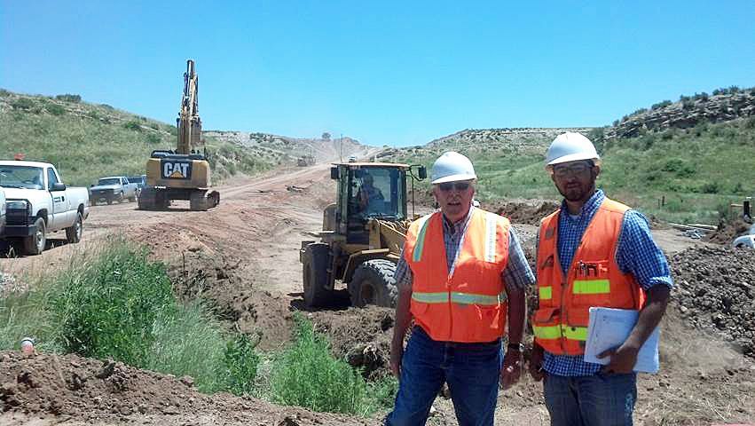 Two workers in orange safety vests at a road construction site with heavy equipment and trucks.