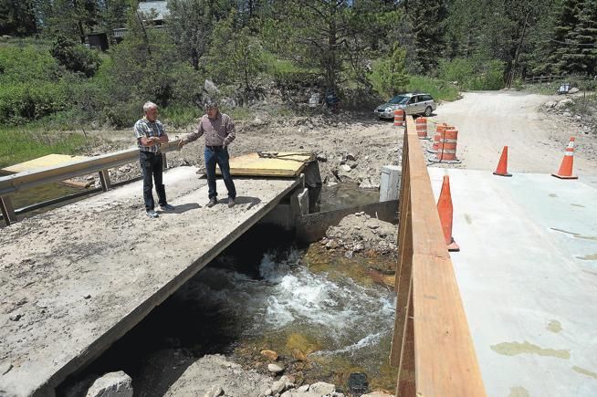 Two people stand on a damaged bridge over rushing water beside a road closure and orange cones