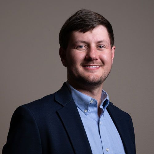 Smiling man in a navy blazer and light blue shirt against a neutral background