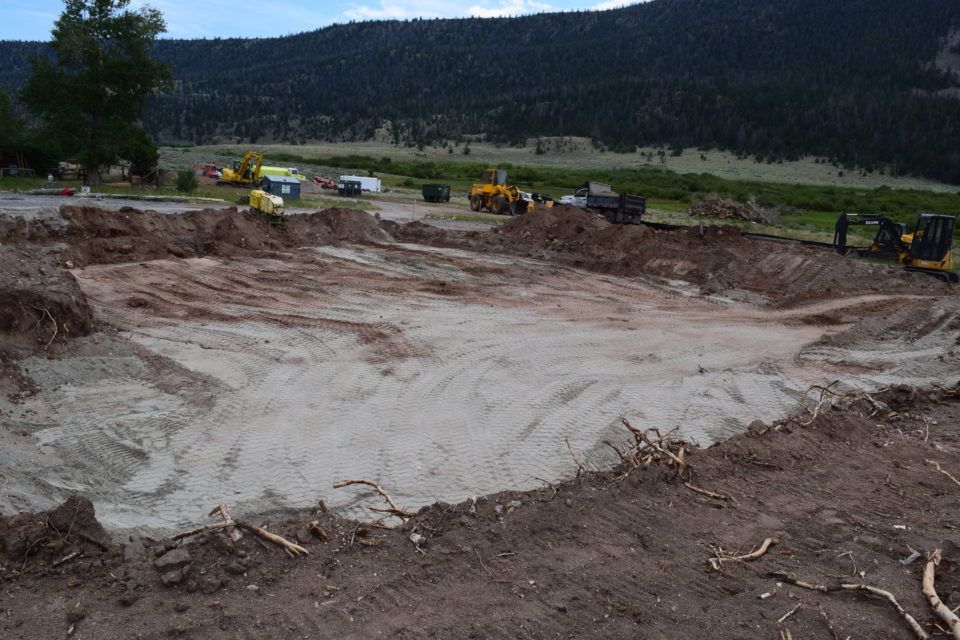 Large excavated dirt pit with construction vehicles in the background and a forested hillside beyond