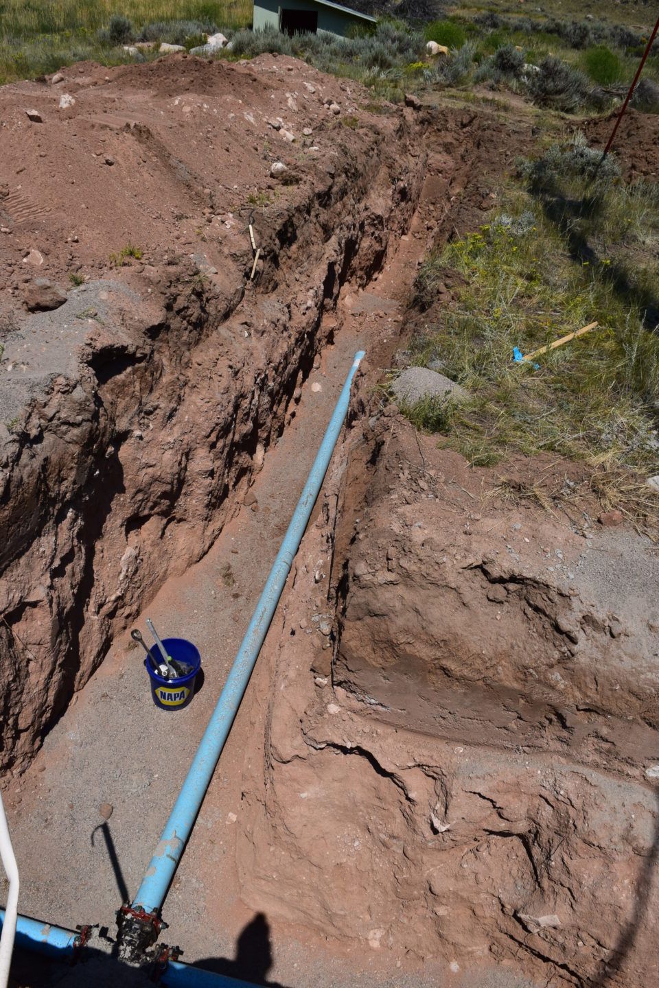 Blue pipe laid in a muddy trench on a dirt hillside, with a tape measure nearby.