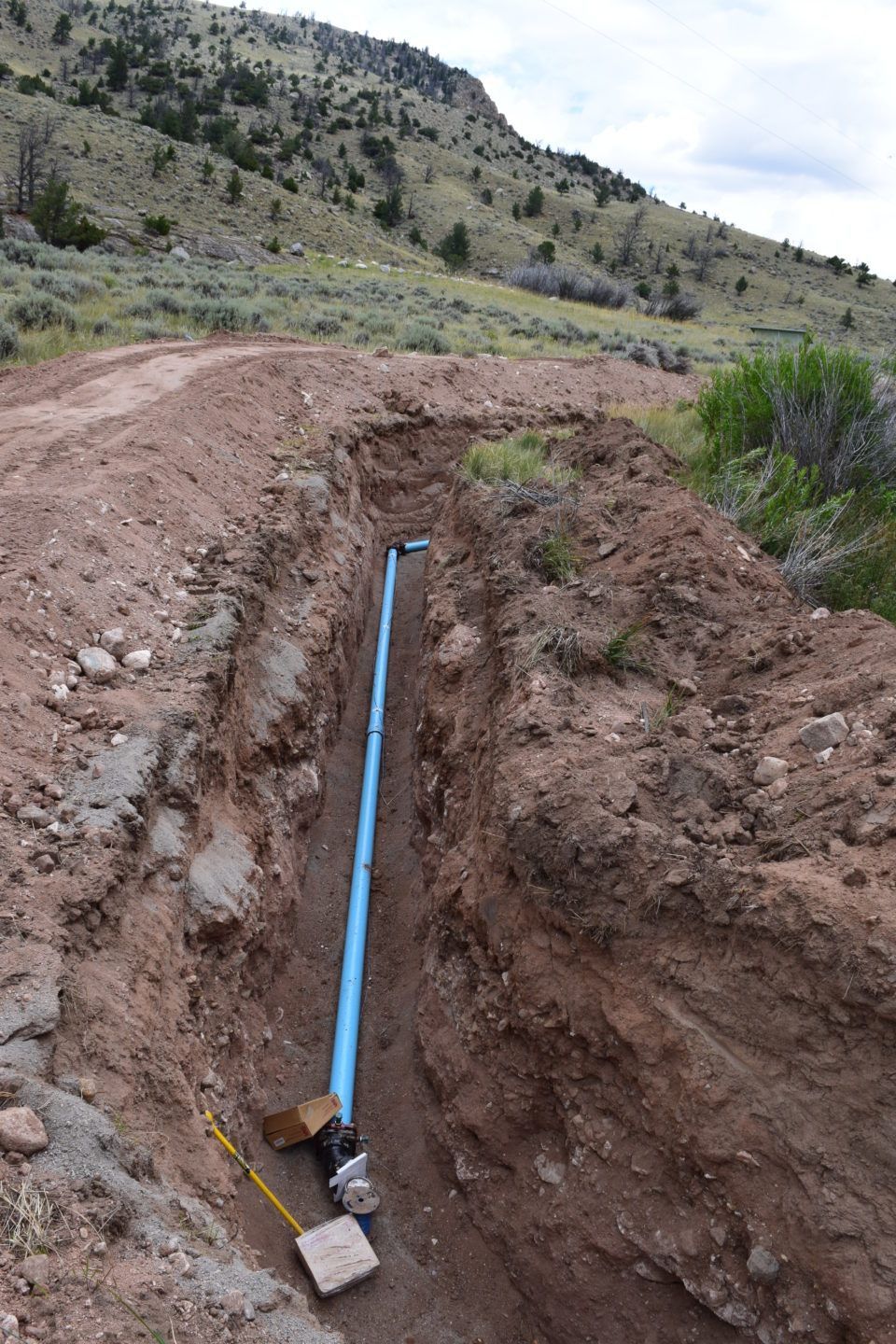 Blue pipe exposed in a muddy trench along a dirt road in a hilly landscape