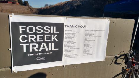 Fossil Creek Trail sign with thank-you notice on a fence, with a bicycle partially visible on the right.