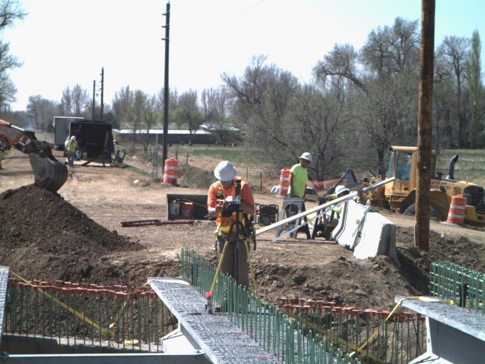 Construction workers near a bridge site, with equipment, cones, and a muddy excavation area under a clear sky.