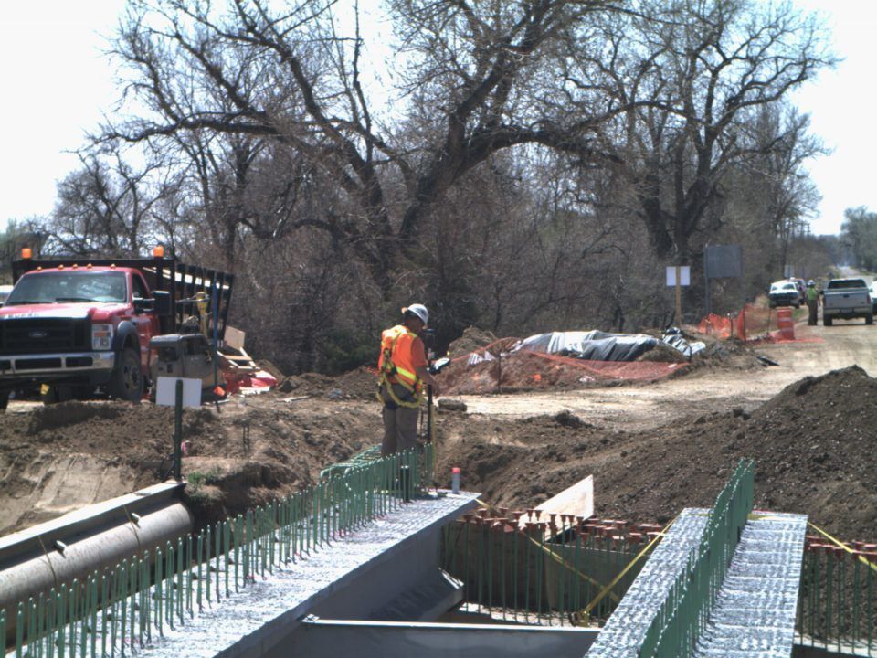 Roadwork crew beside a damaged bridge over a muddy ditch, with trucks and construction barriers nearby