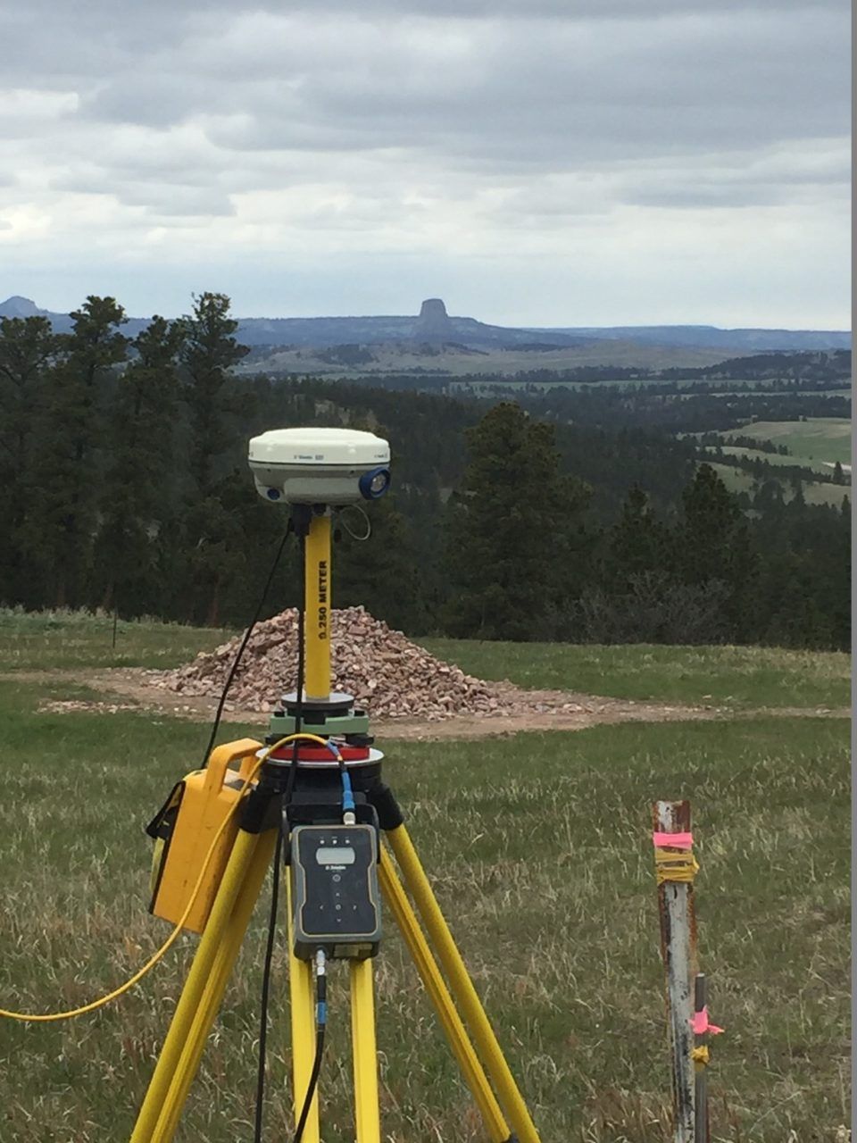 Survey equipment on yellow tripod in grassy field with distant hills and cloudy sky