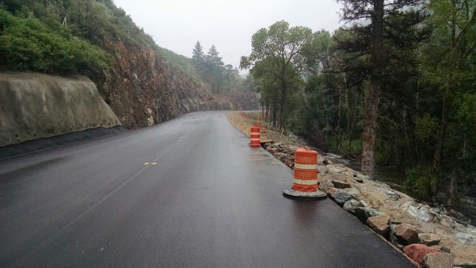 Wet mountain road with orange traffic barrels beside a rocky, tree-lined shoulder