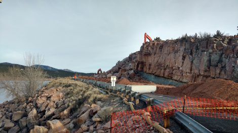Rocky cliffside construction site with red barriers, excavator, and a dirt road