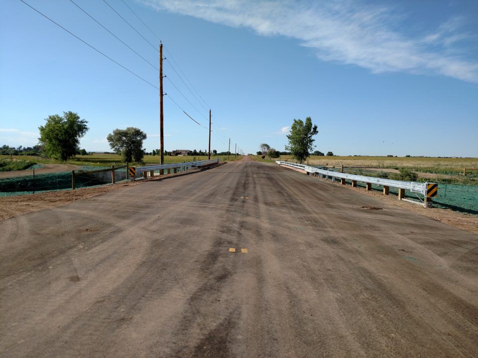 Empty rural road under a clear blue sky, with utility poles and guardrails along flat farmland.