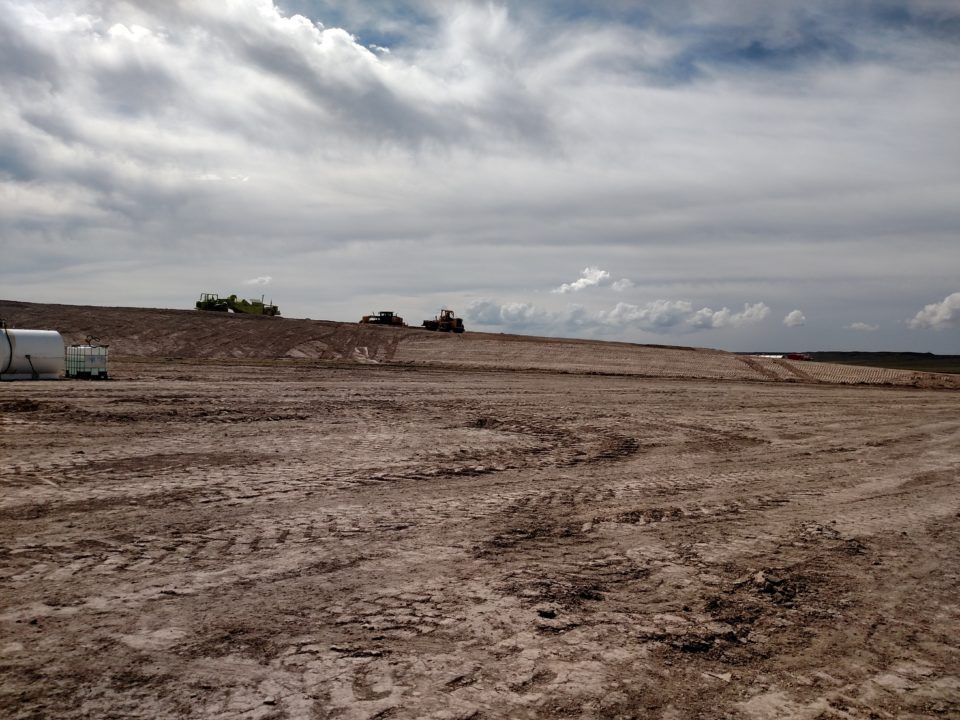 Wide muddy dirt field under a cloudy sky, with a low embankment and distant trees on the horizon.
