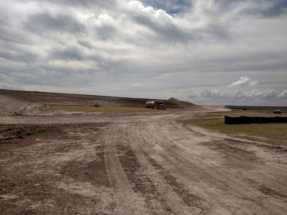 Wide dirt road across a barren landscape under a cloudy sky, with a small red vehicle in the distance.