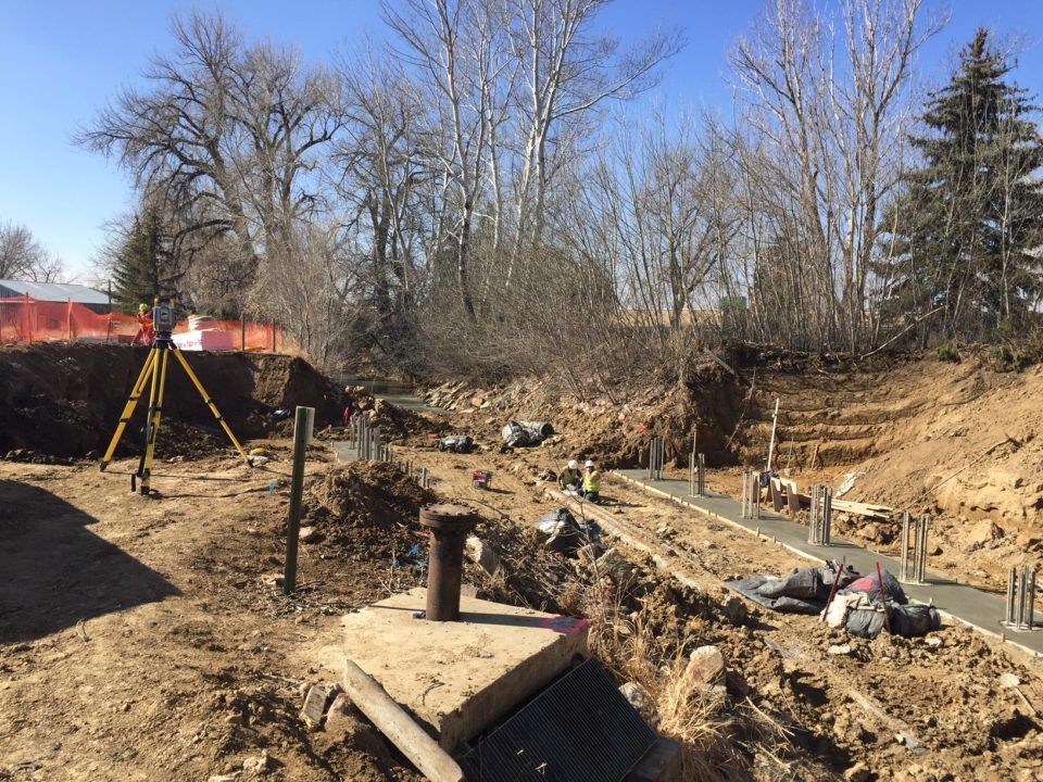 Construction site with trench, survey tripod, and exposed pipes beside a red bridge under bare trees