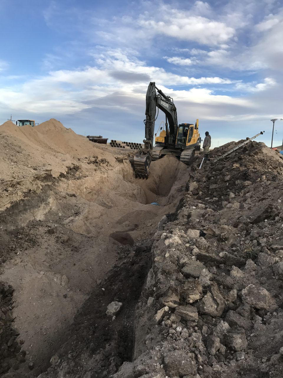 Excavator digging a trench between dirt mounds under a cloudy sky