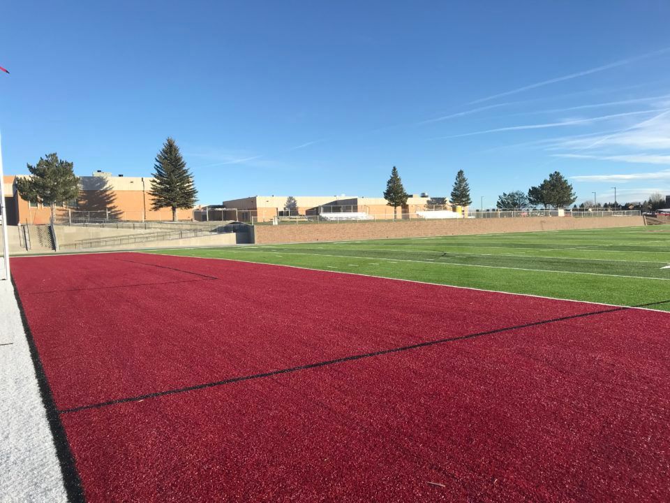 Red running track beside a green sports field under a clear blue sky, with buildings in the distance