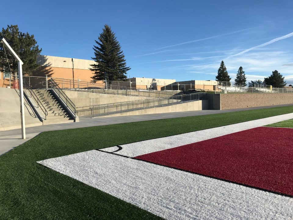 School track field with red turf, white sideline, bleachers, stairs, and buildings under a clear sky