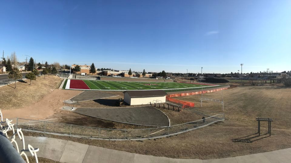 Wide outdoor sports field with running track, dirt infield, and bleachers under a clear blue sky