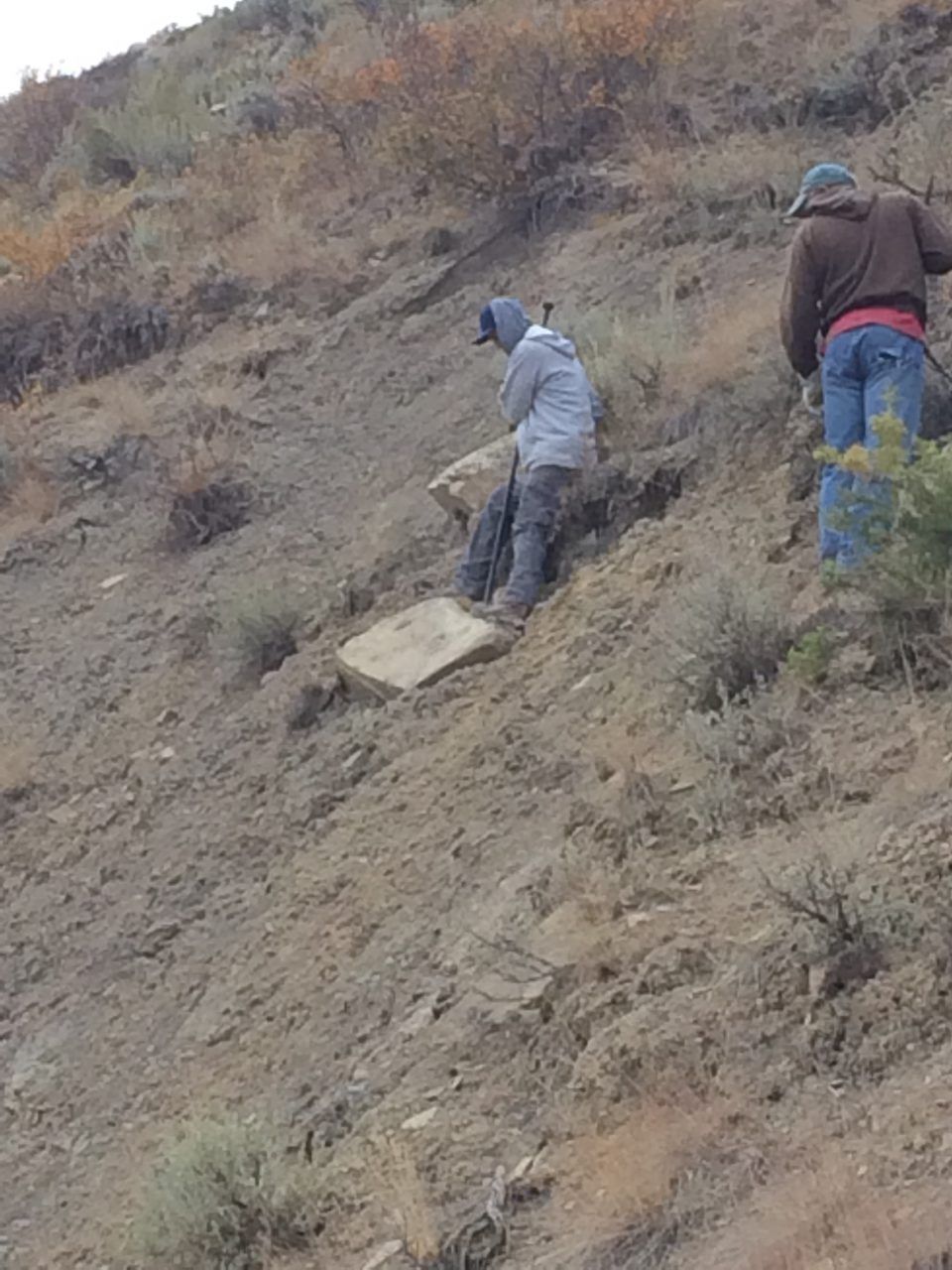 Two people hike a steep dirt slope, one carrying a white board or rock.